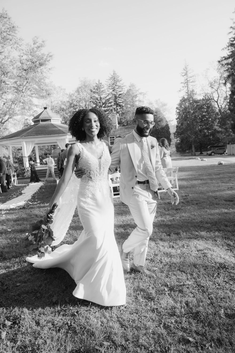 A couple walking together outdoors in elegant white wedding attire near a garden gazebo.
