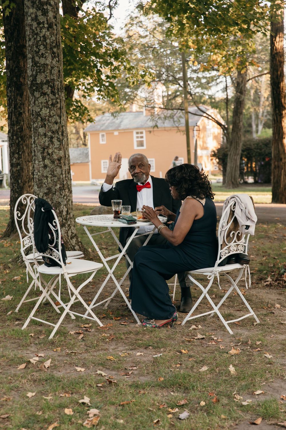Two people enjoying tea at a small white bistro table set outdoors under fall trees.