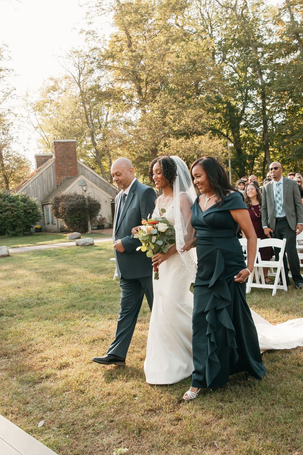 A bride walks down a garden path with family members during an outdoor wedding ceremony.
