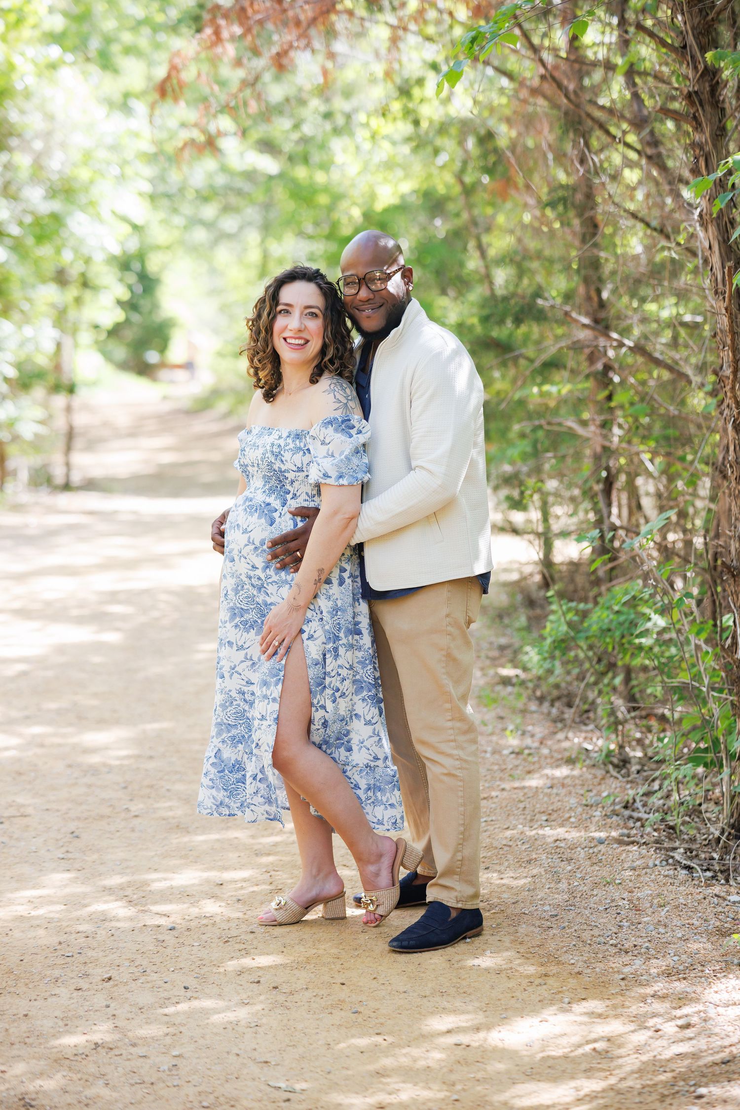 A couple shares a romantic moment together on a sunlit forest path during their maternity photoshoot in Dallas