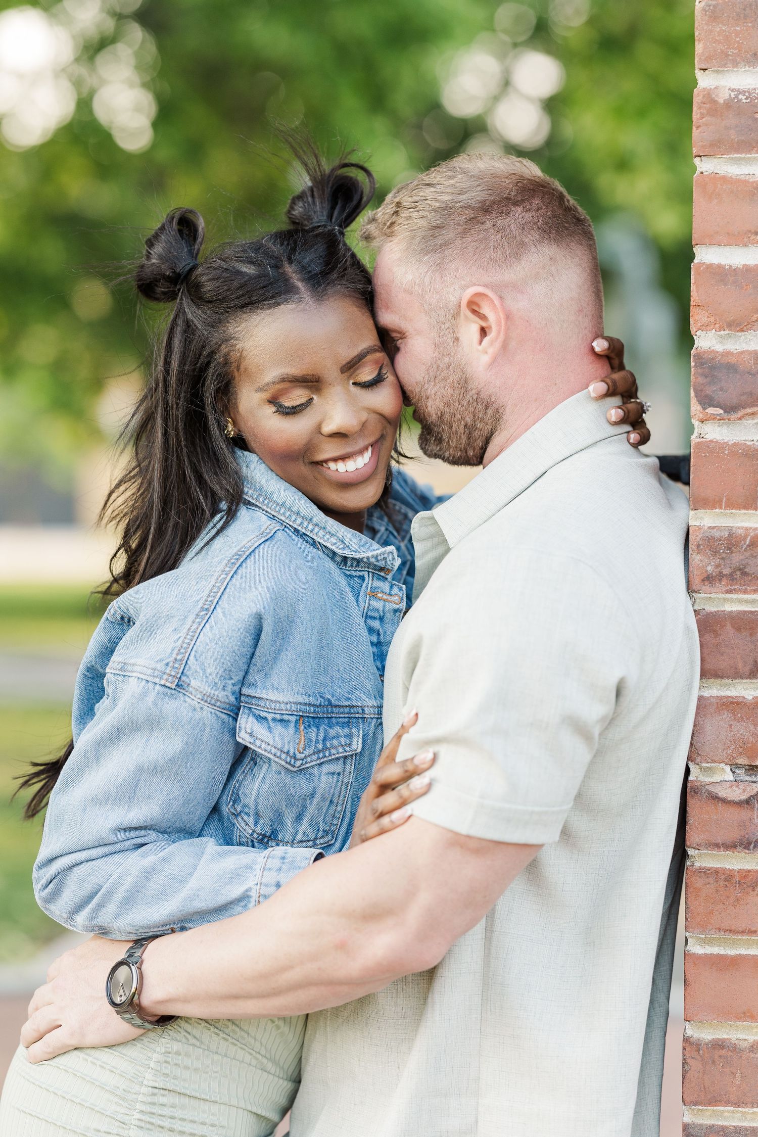 A couple shares an intimate embrace against a brick wall in the park, dressed in denim and white casual attire.