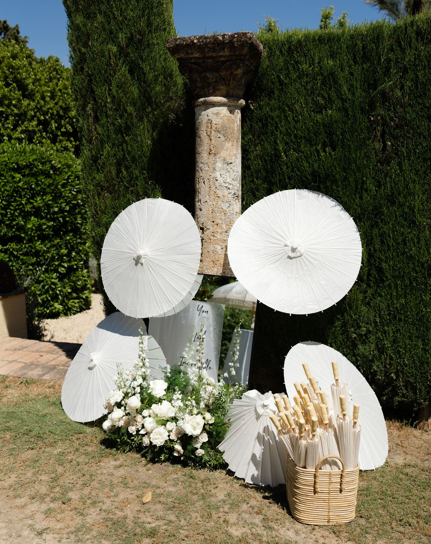 White paper parasols and floral arrangements artfully displayed against green cypress trees in an elegant garden setting.