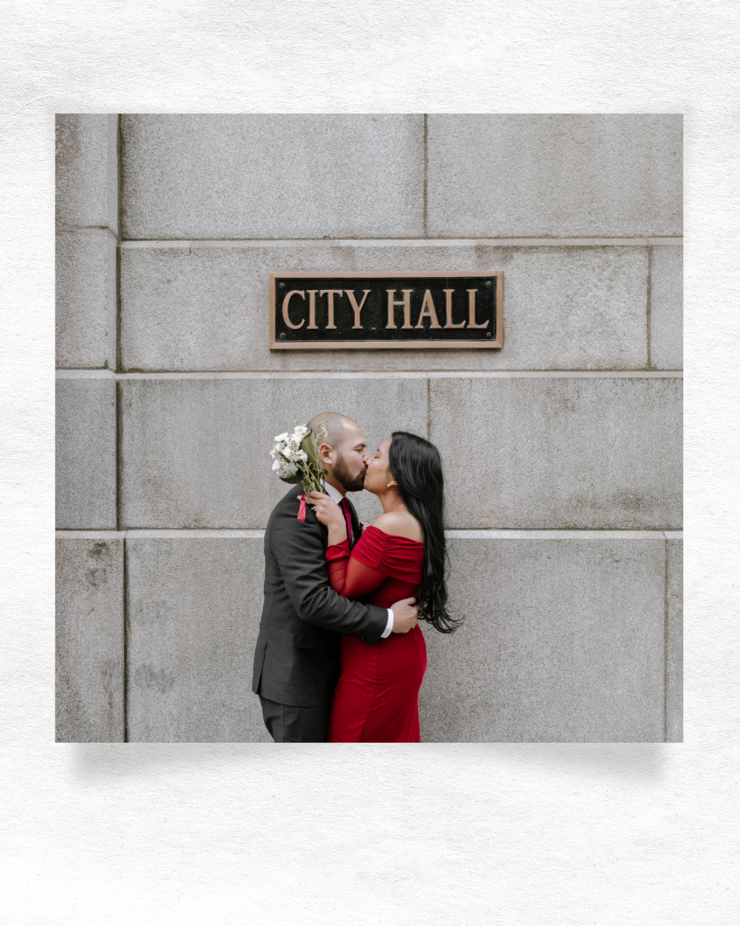 A romantic couple embraces outside City Hall, with the bride wearing a red dress and holding white flowers.