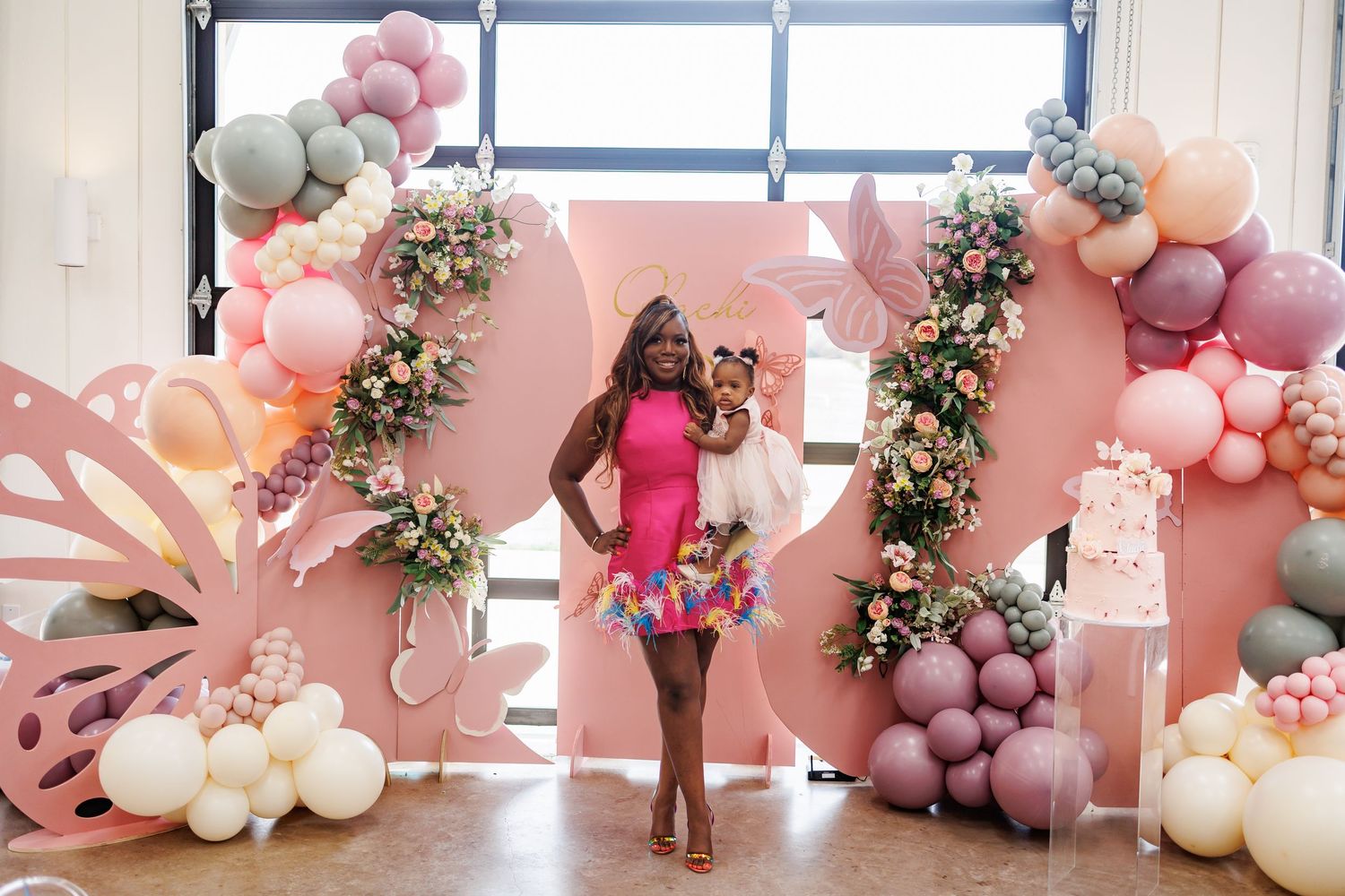 Mom and birthday baby stand in front of a balloon arch installation in pink and white with floral accents at a Bella Cavalli first birthday