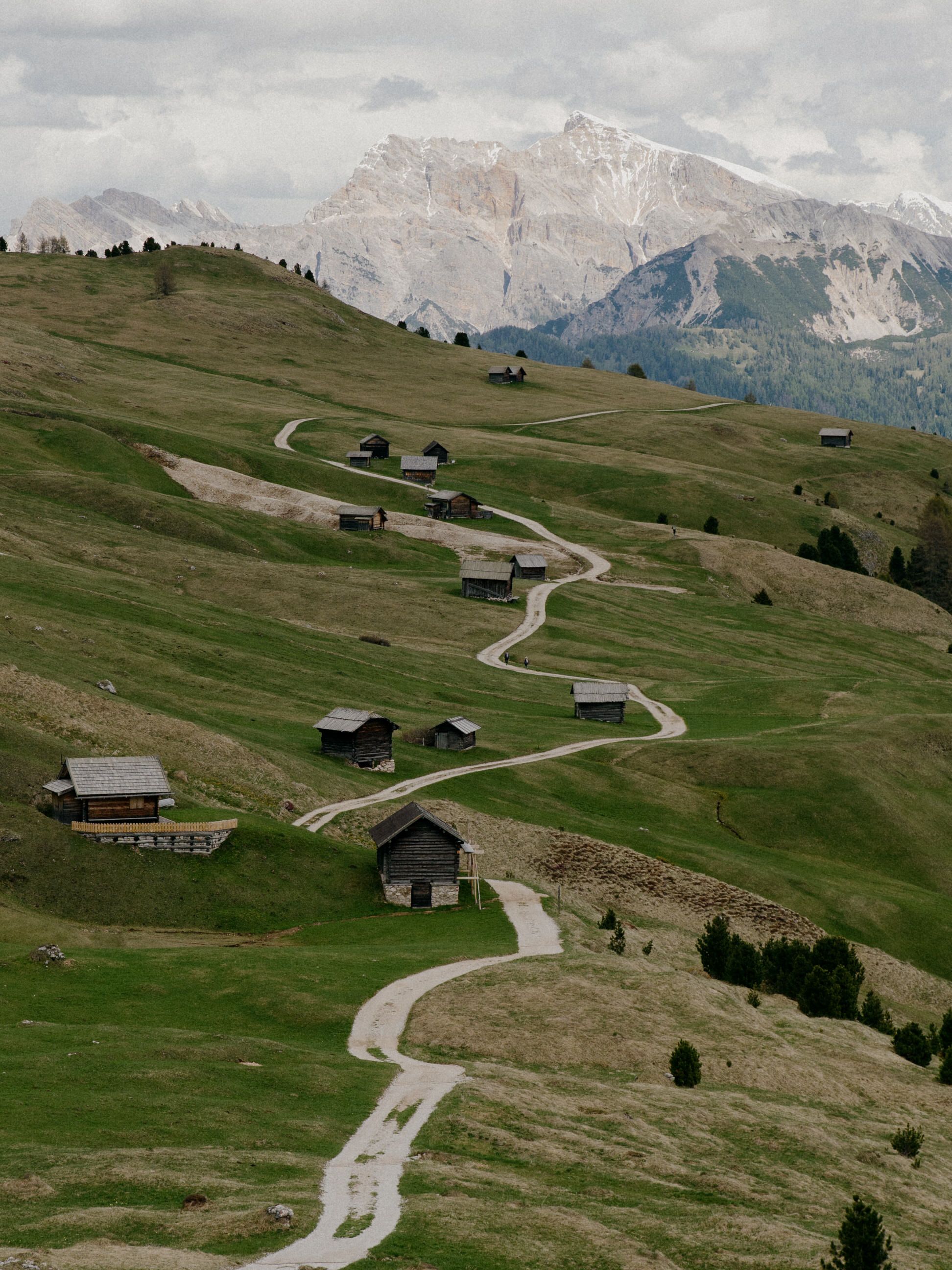 A winding mountain road cuts through green alpine meadows with wooden chalets and dramatic Dolomite peaks in the background.