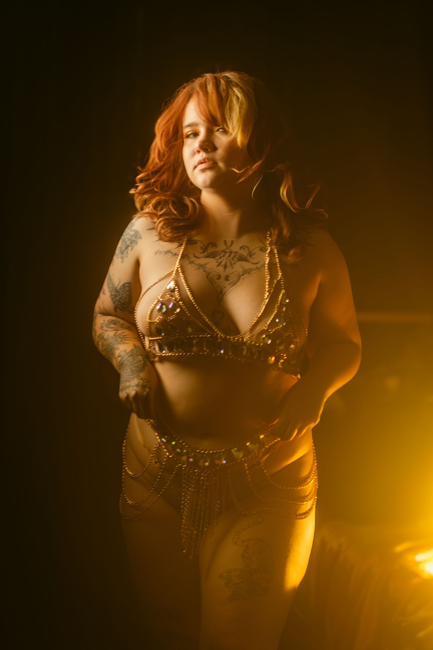 Dramatic golden-lit boudoir photo of figure in studded bralette with tattoos against dark background with atmospheric lighting.