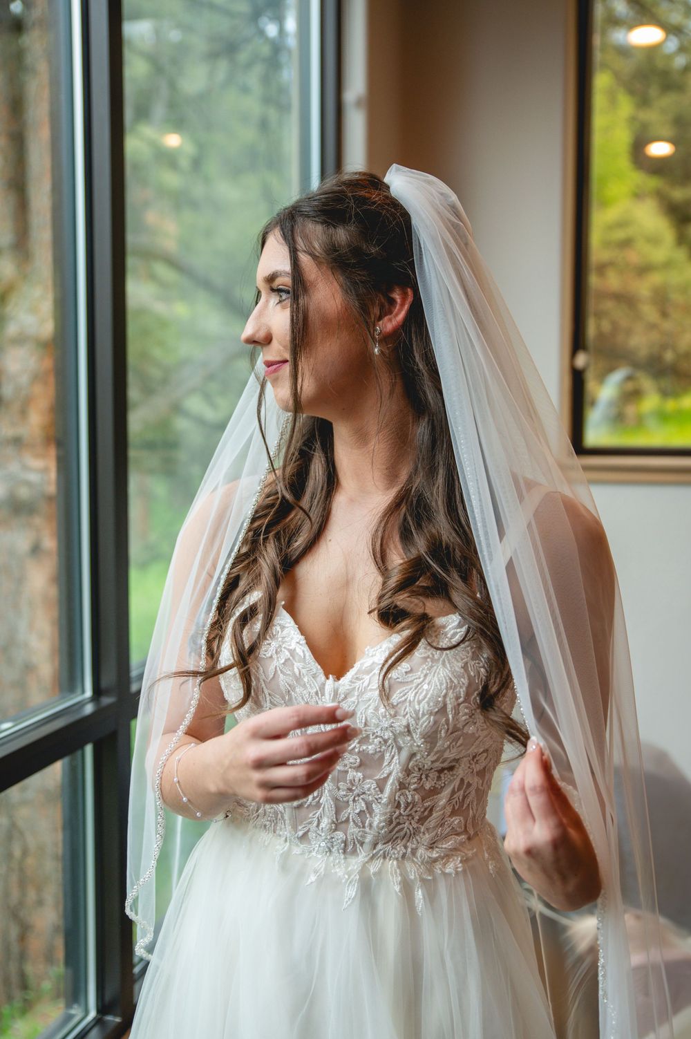 A bride in a lace wedding gown and veil gazes thoughtfully out a window in soft natural light.