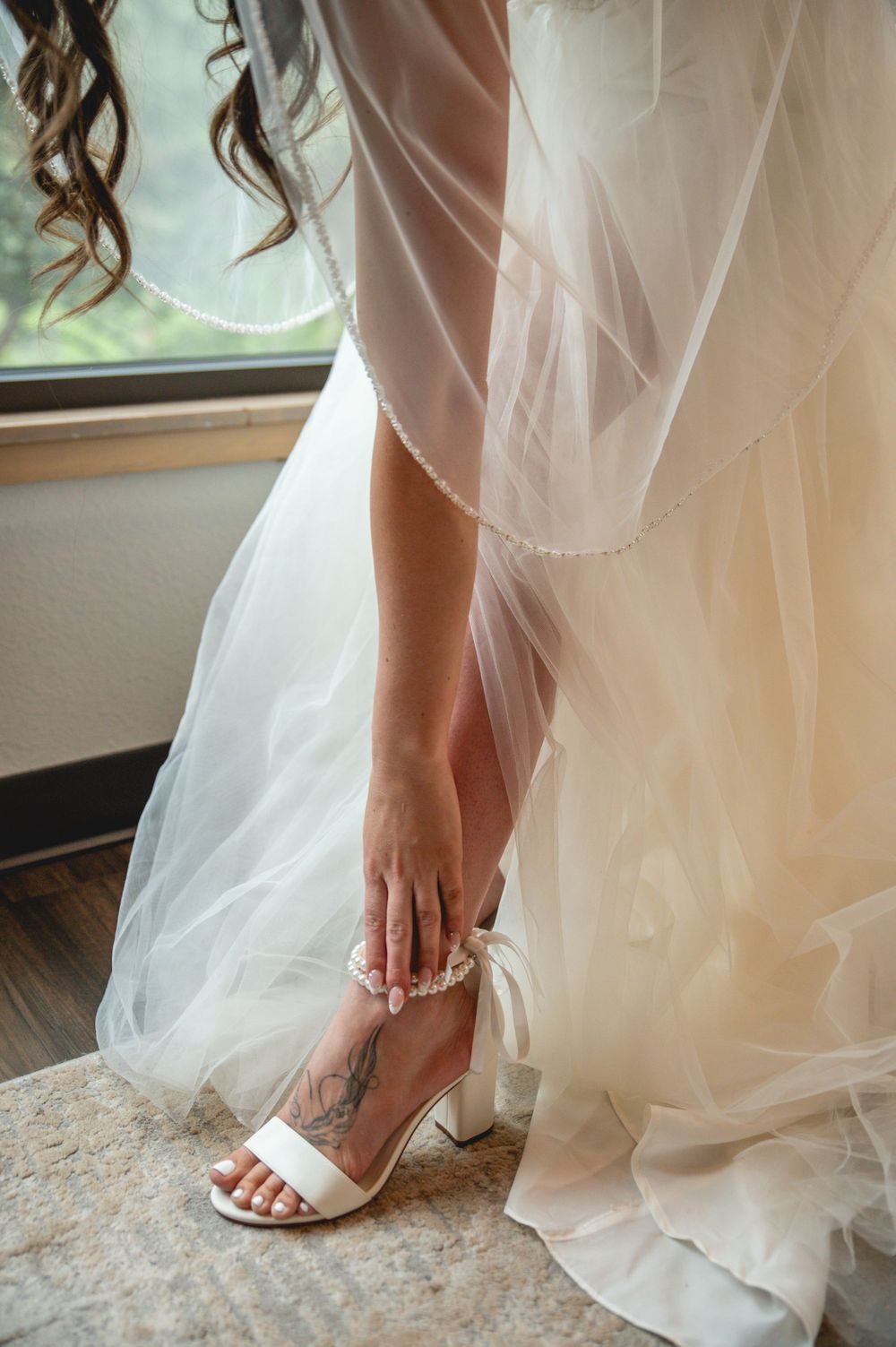 A bride's foot with delicate strappy white sandals and ankle tattoo shown beneath flowing tulle wedding dress layers.
