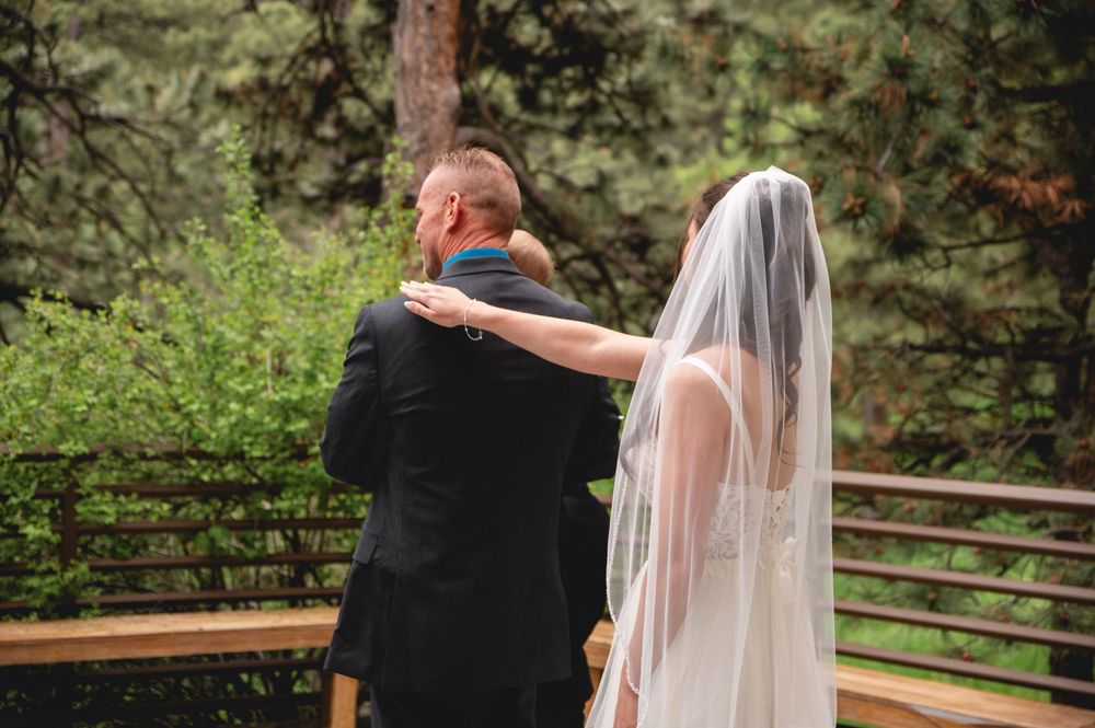 An emotional moment captured from behind as a bride in her veil embraces someone wearing black at an outdoor wedding ceremony.