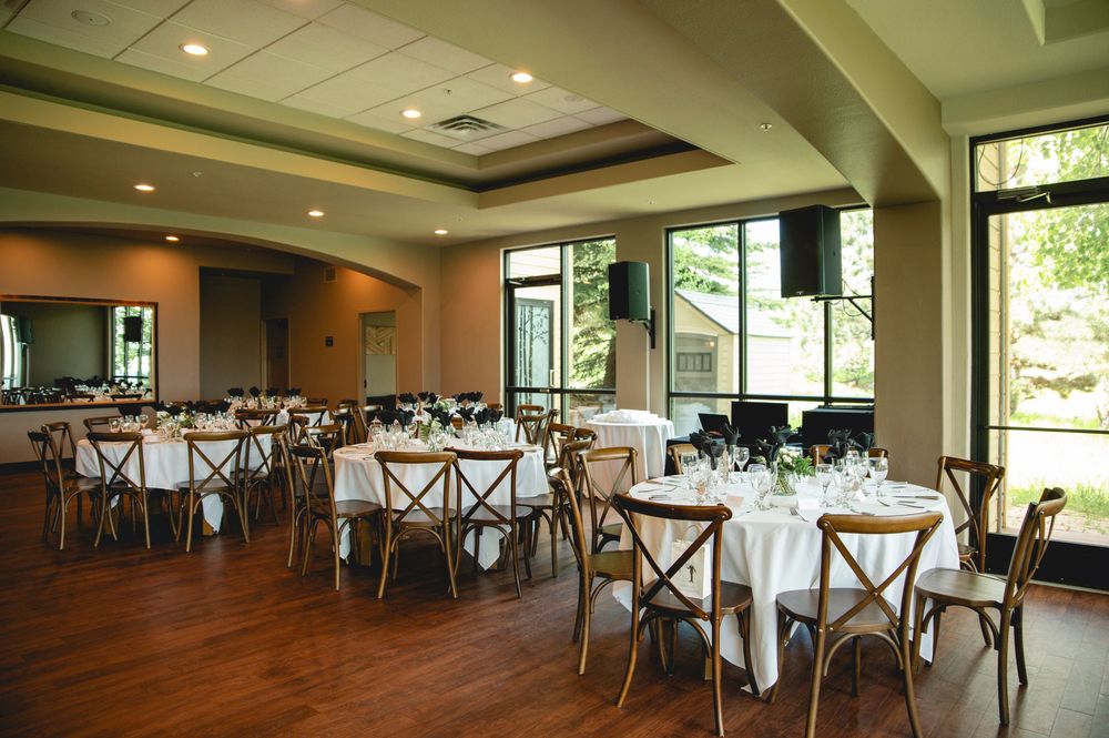 An elegant banquet hall with hardwood floors, windows, and round tables set with white linens and cross-back chairs.