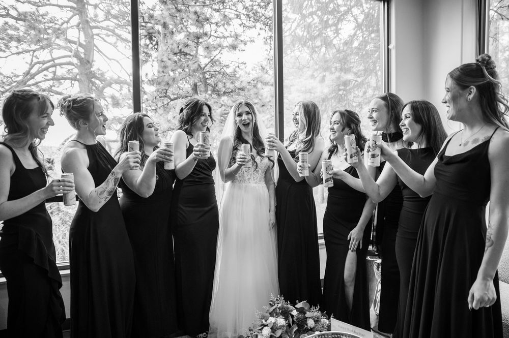 Wedding party in black dresses stands in a line against bright windows creating a dramatic silhouette effect.