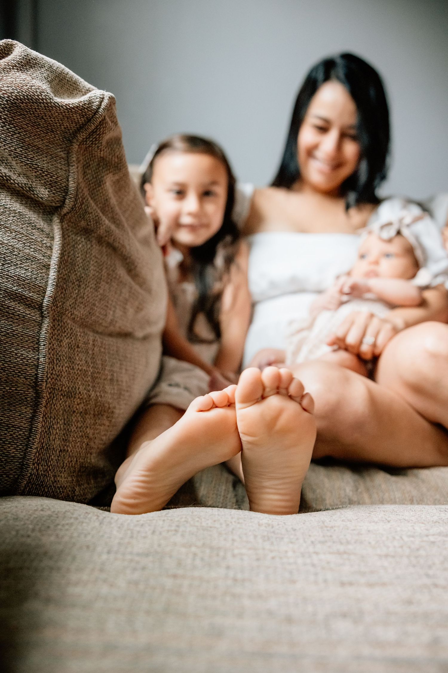 A close-up of bare feet on a gray couch while family members relax together at home.