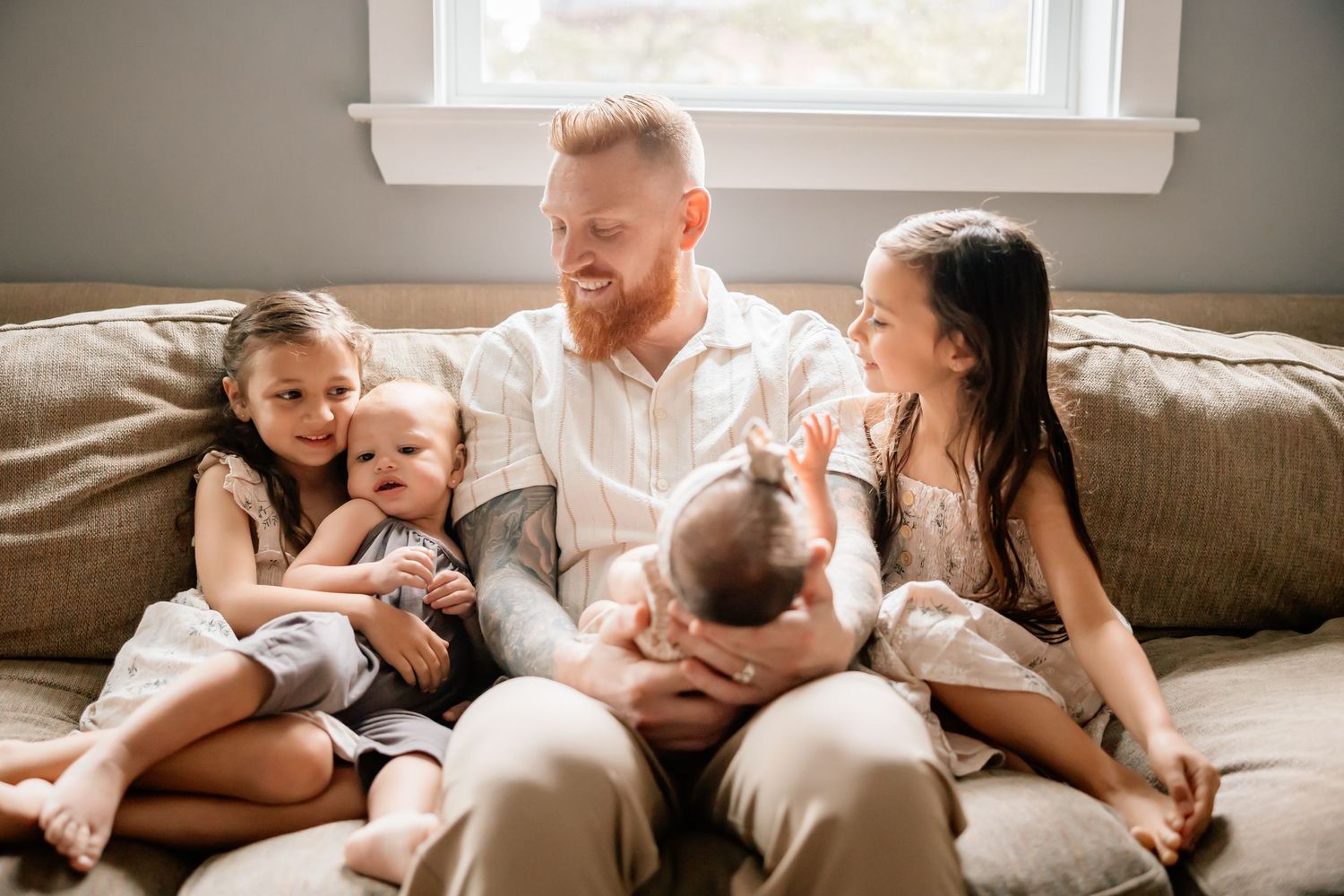 A parent sits on a beige couch with four young children in a sunlit living room.