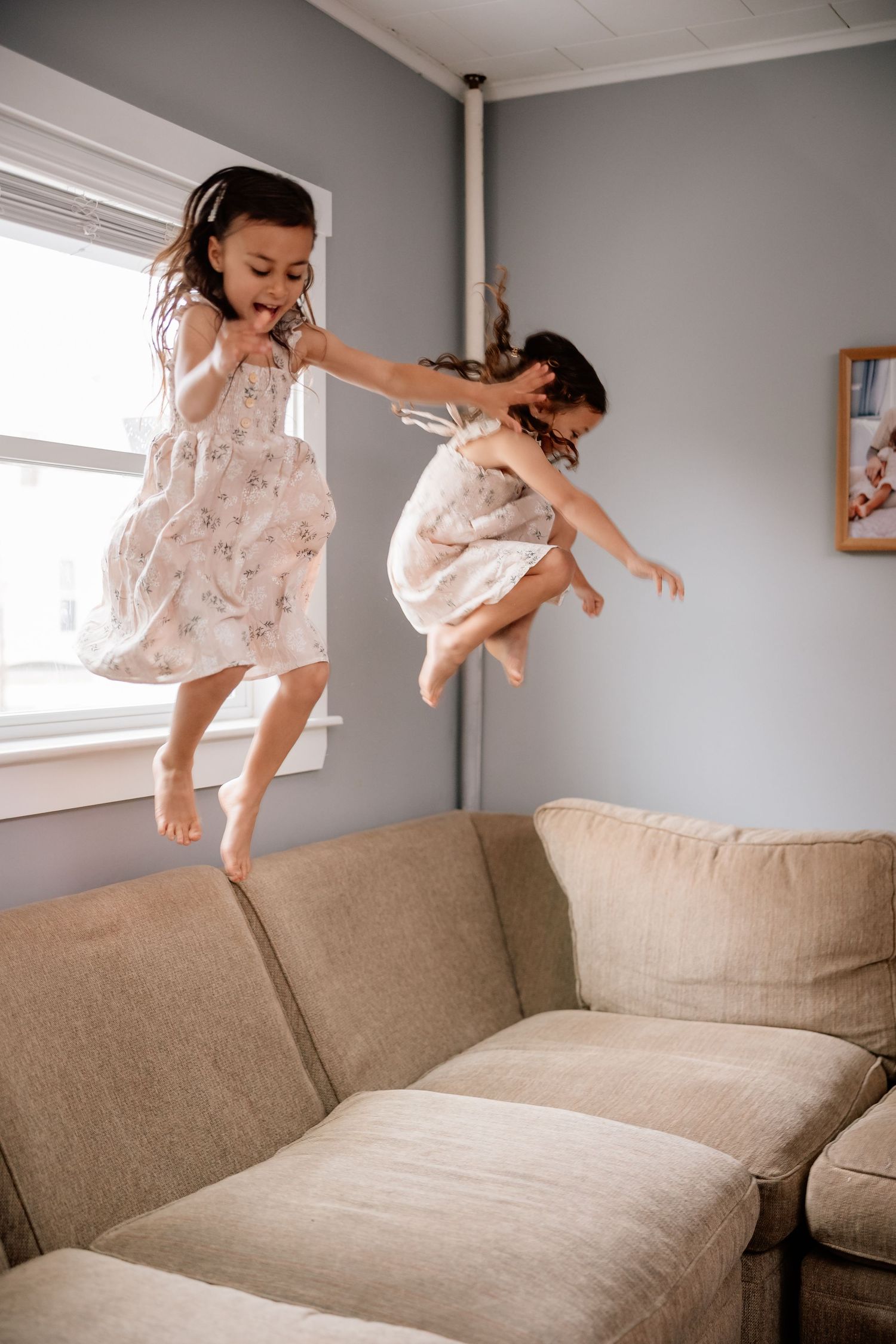 Two young girls in matching floral dresses jumping excitedly on a beige couch in a gray-walled room with natural light.
