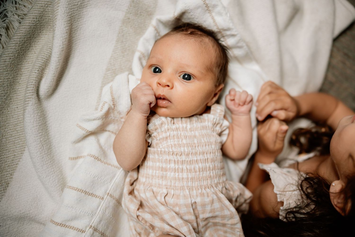 Small baby wearing white smocked romper lies on cream colored blanket looking up with alert expression.