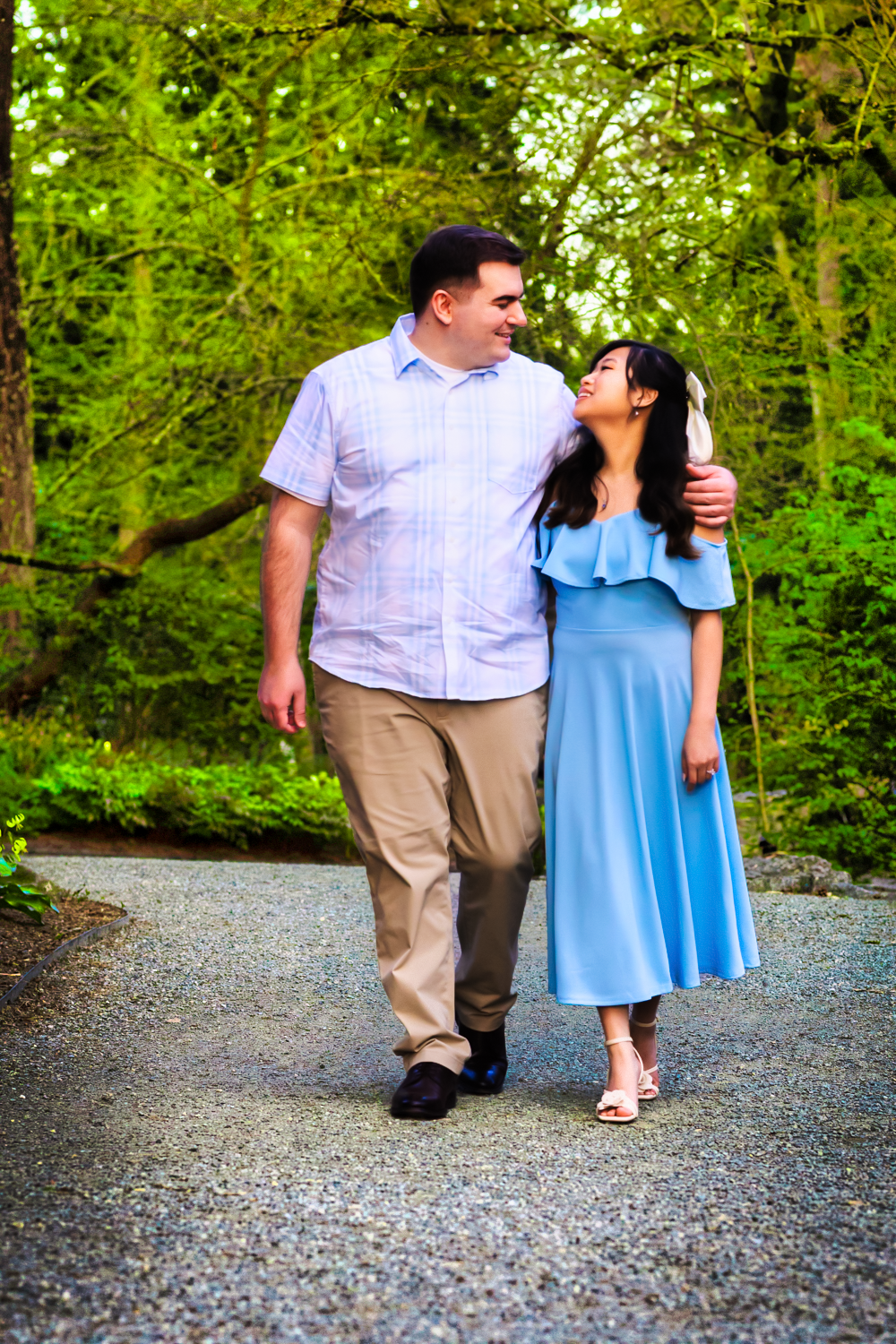 A couple walks along a gravel path surrounded by green trees while sharing a loving glance.