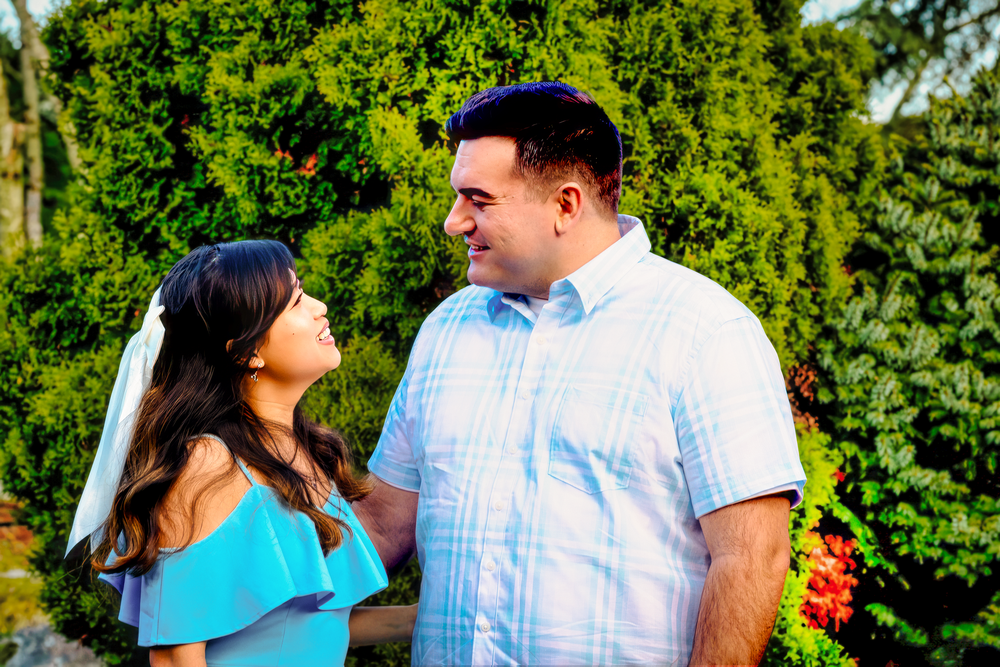 A couple wearing turquoise blue and white outfits smile at each other in front of lush green garden foliage.