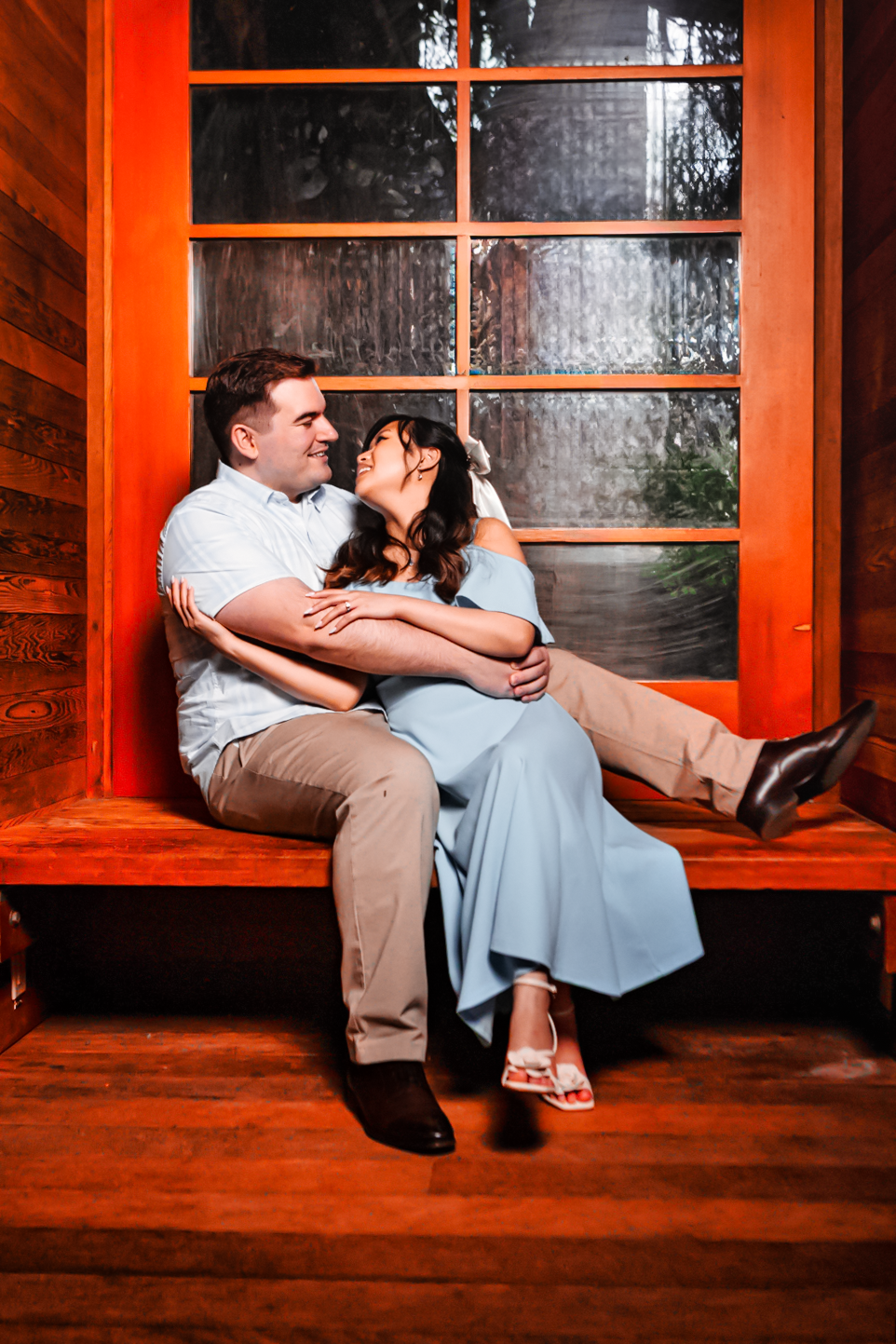 A couple shares an intimate moment sitting in a vibrant orange window frame on a rustic wooden porch.