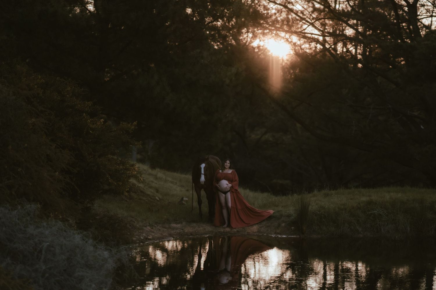 Silhouetted couple stands by a tranquil pond at sunset with tall trees creating a moody forest backdrop in Mansfield, Victoria