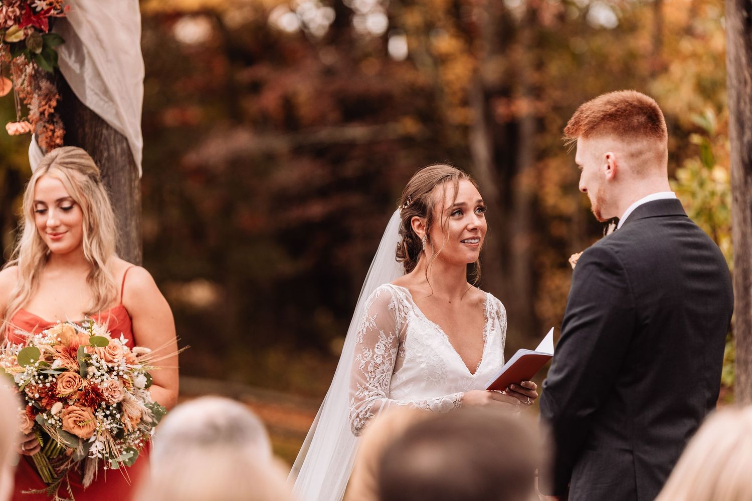 A couple exchanges vows during an outdoor autumn wedding ceremony while a bridesmaid stands nearby holding a bouquet.