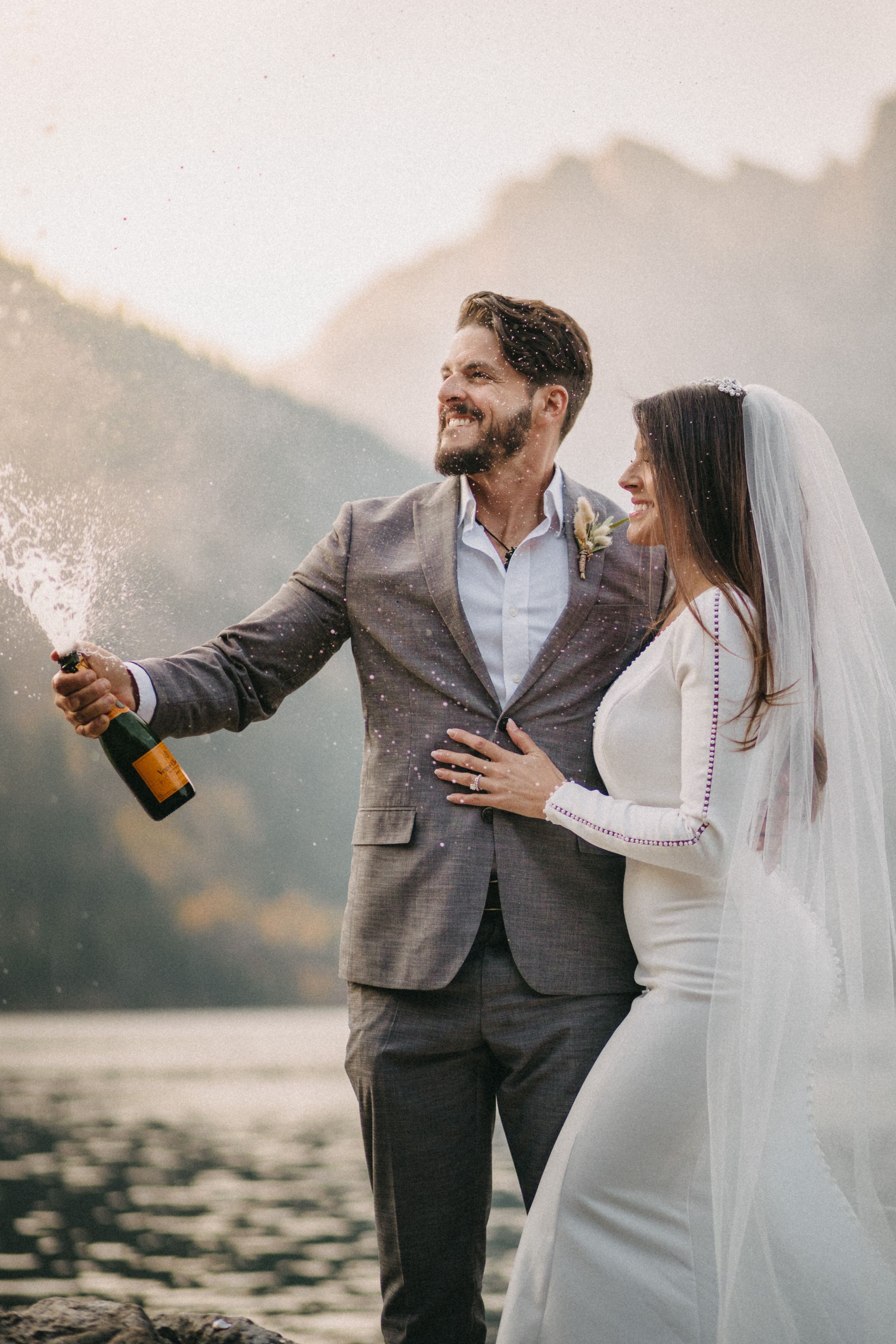Newlywed couple celebrates with champagne by a mountain lake on their wedding day during sunset.