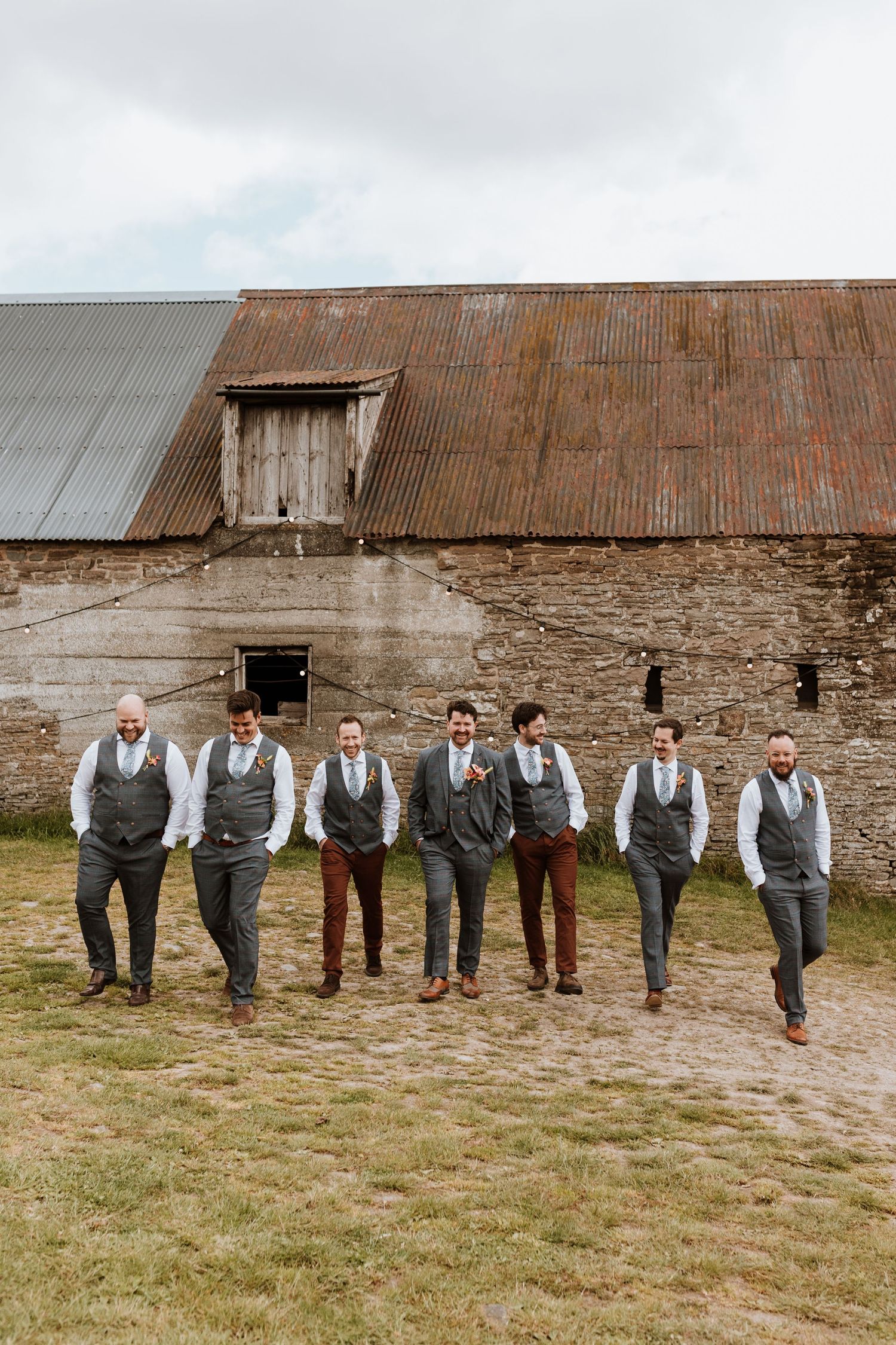 A line of people in wedding attire lean against a rustic barn wall wearing gray vests and dress pants.