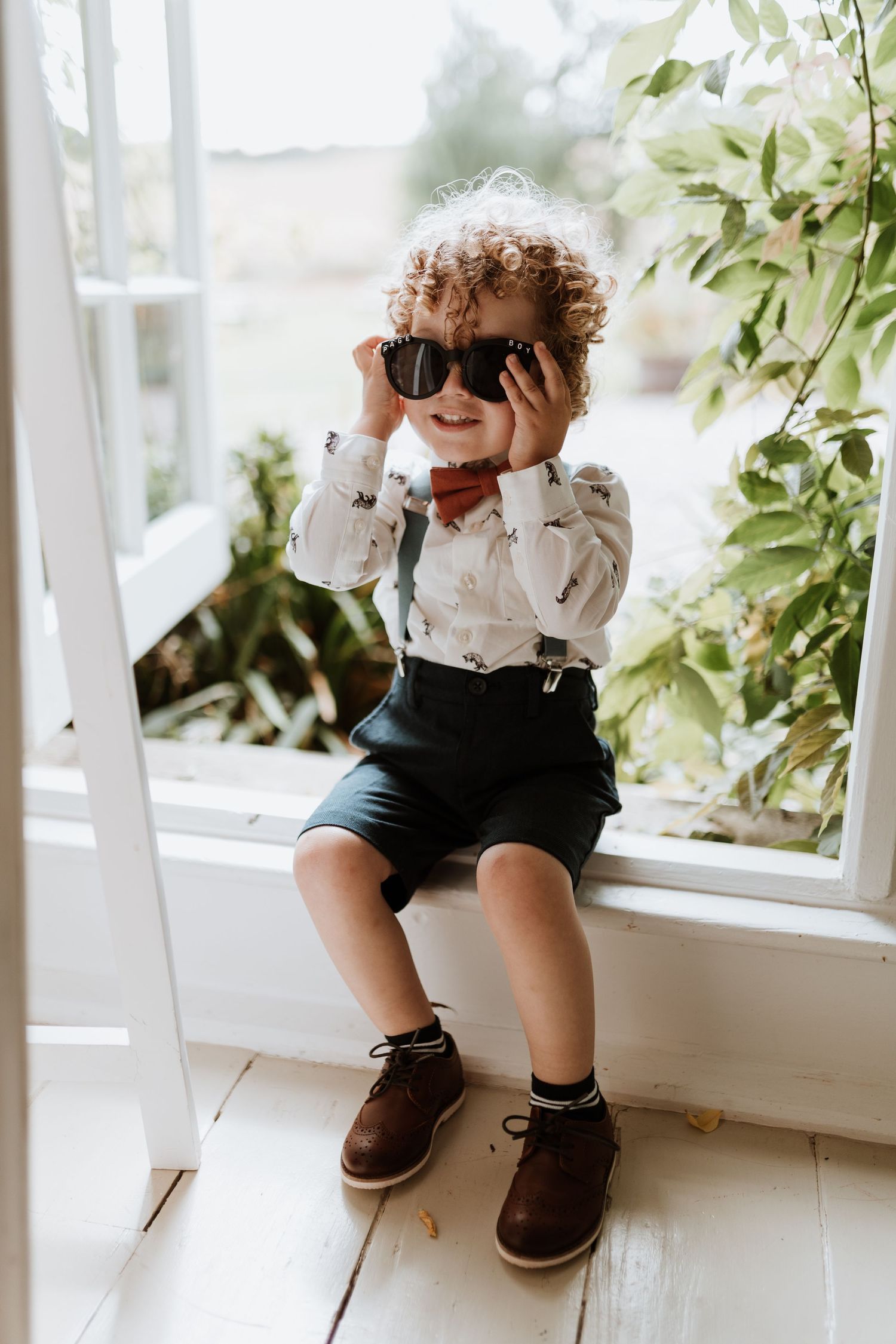 A small child wearing suspender shorts and a bow tie sits by a window, playfully holding dark sunglasses to their face.