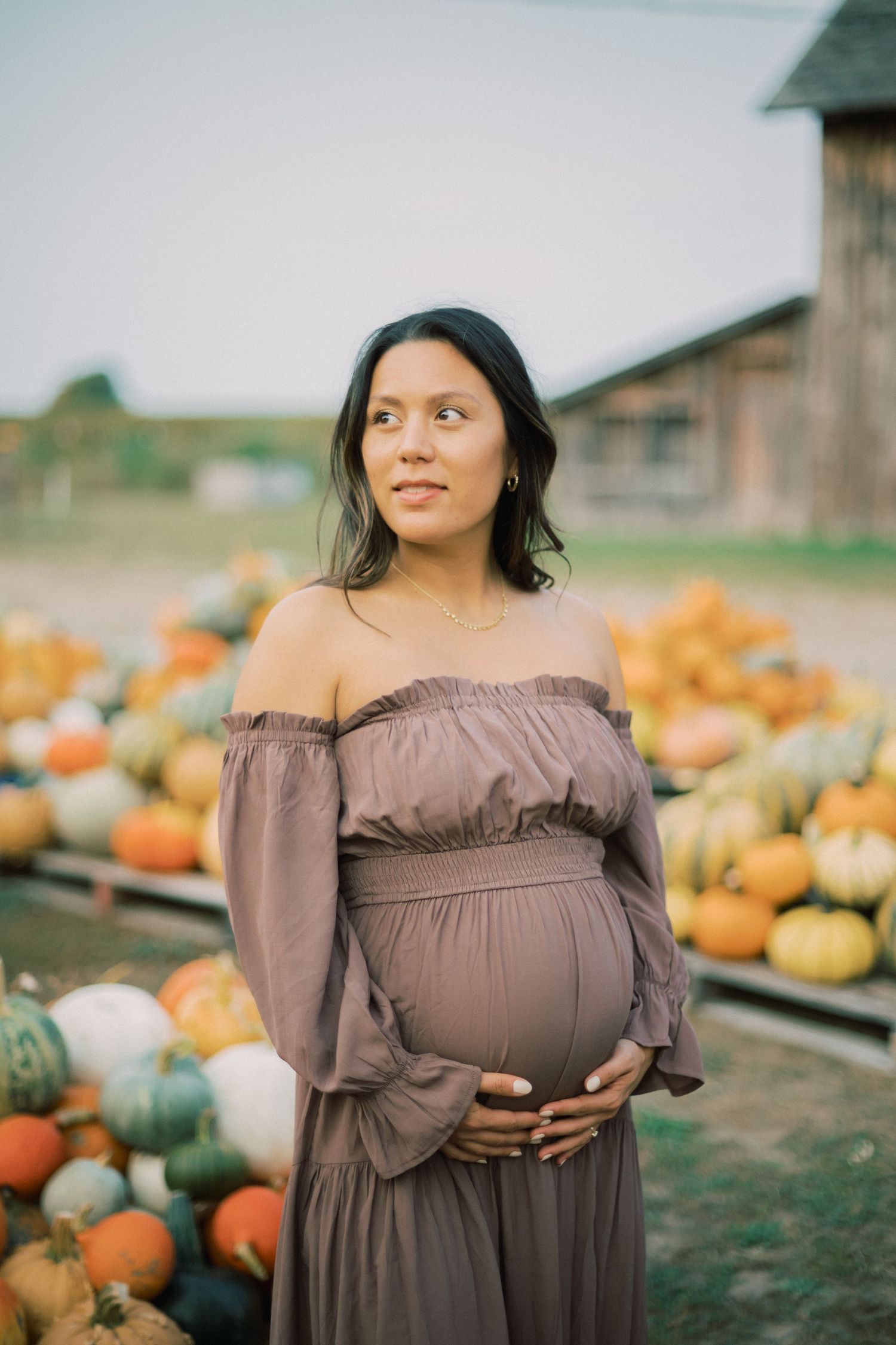 A maternity portrait at a pumpkin patch with gourds and farm buildings in the background.