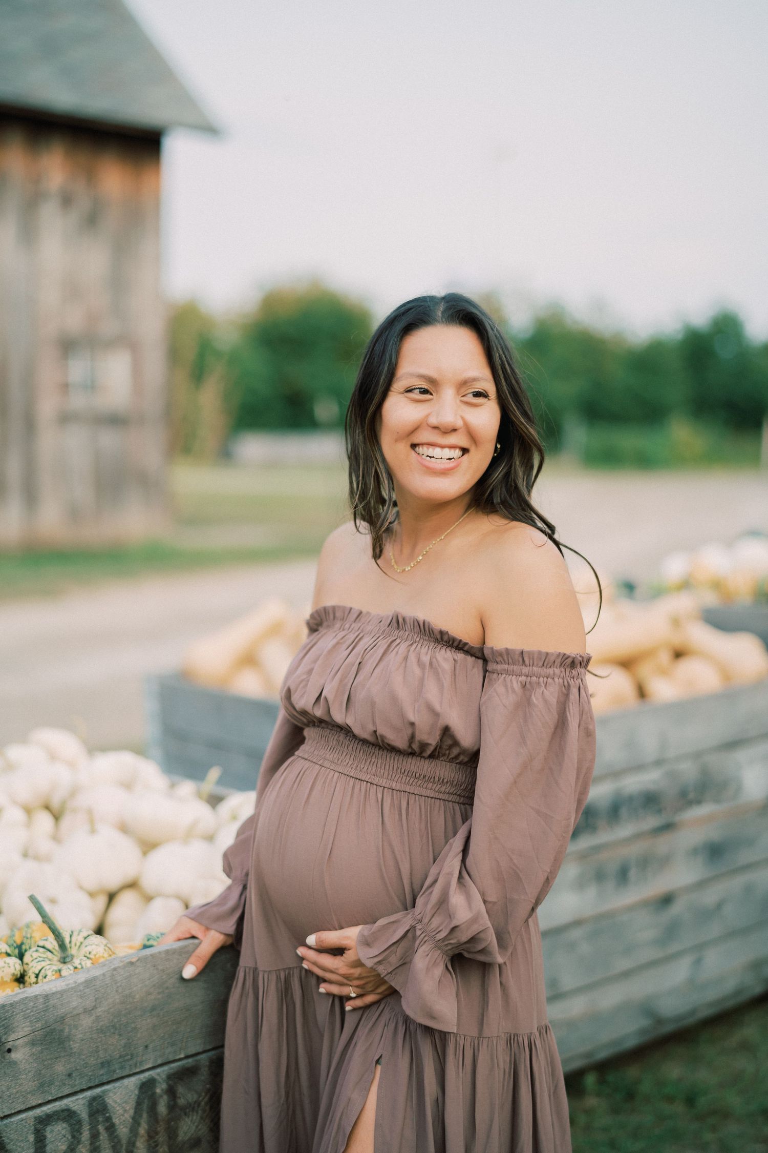Smiling pregnant woman in brown dress standing beside wooden farm crates.