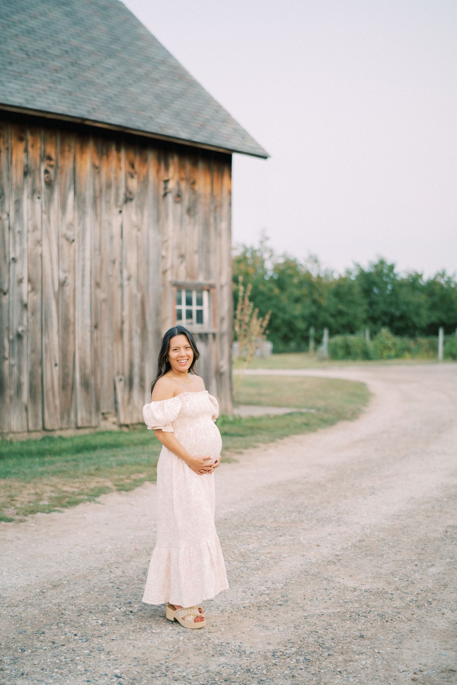 A soft pink dress stands out against the weathered wood of a rustic barn on a country path.
