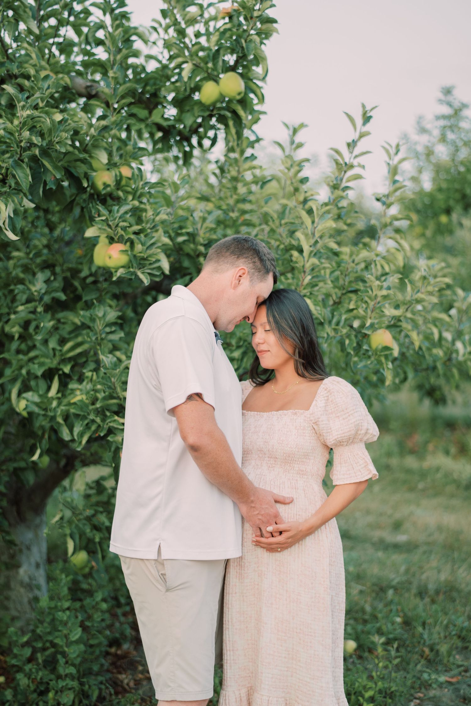 A couple shares an intimate moment in a sunlit apple orchard wearing light colored clothing.