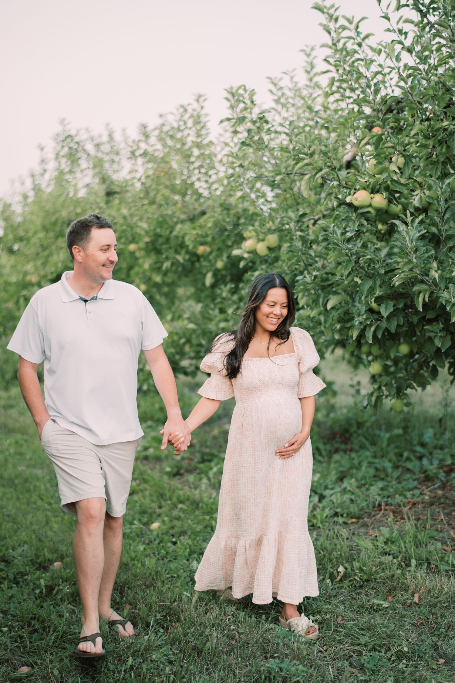 A couple walks hand in hand through an orchard wearing coordinated light-colored outfits.