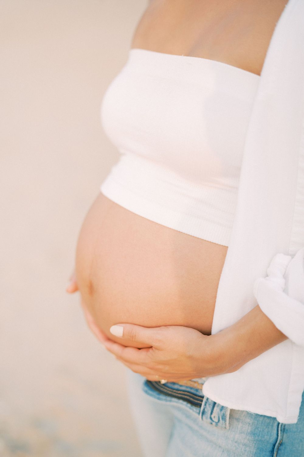 Close-up view of pregnant belly in white crop top and jeans against light background.