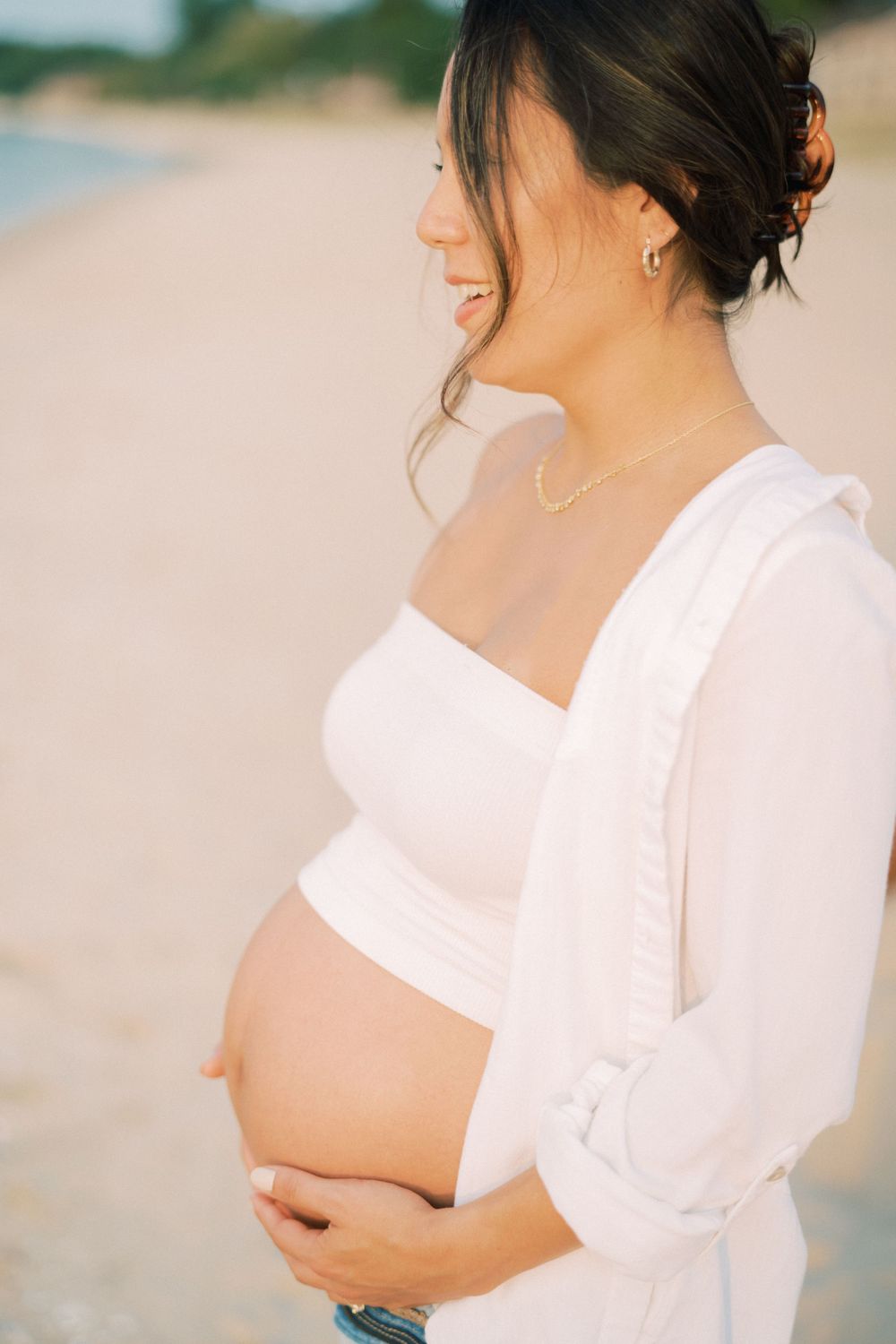 Side view of pregnant woman in white top smiling on beach at sunset.