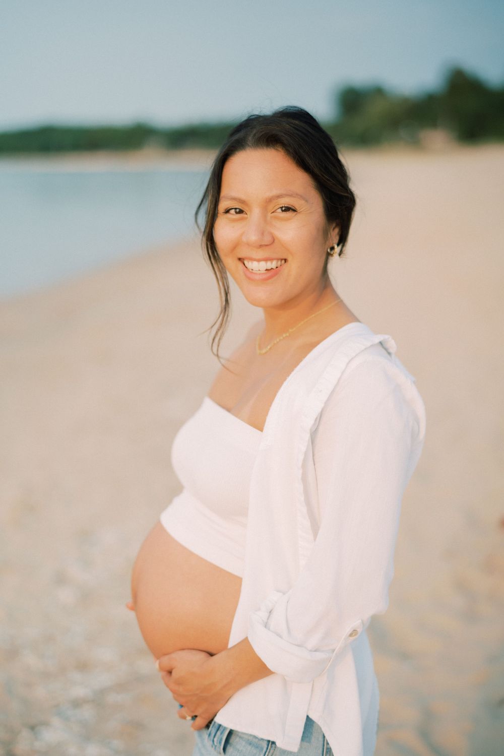 Expectant mother in white top and jeans smiles on sunny beach.