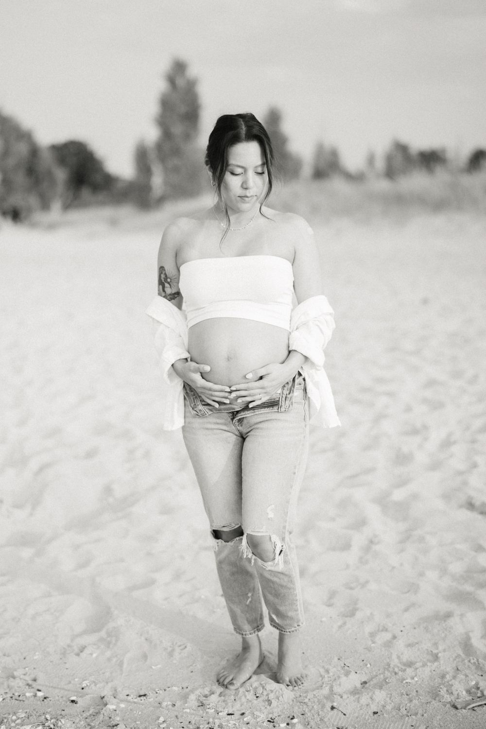 Black and white maternity photo of pregnant woman in casual attire standing on beach.