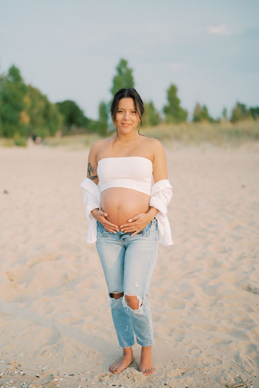 Expecting mother in casual beach attire stands barefoot on sandy beach during pregnancy photoshoot.
