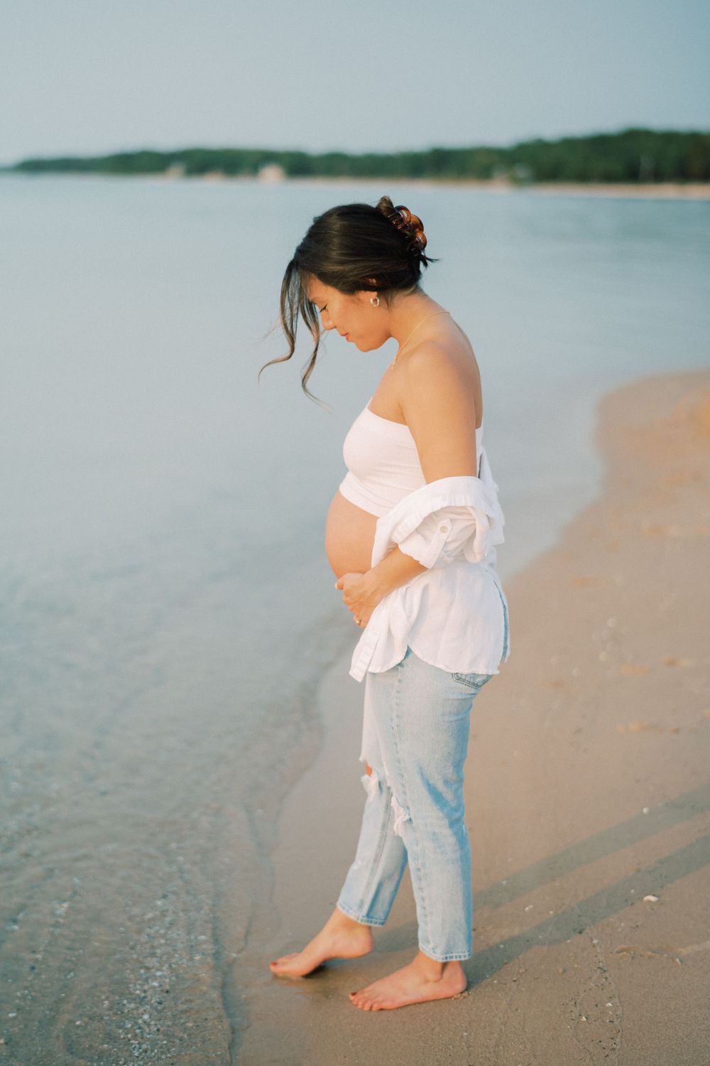 Pregnant woman in white top and distressed jeans walks along shoreline at sunset.