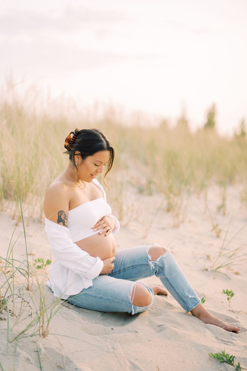 A maternity photo shows a pregnant woman in distressed denim sitting on sandy dunes at sunset.