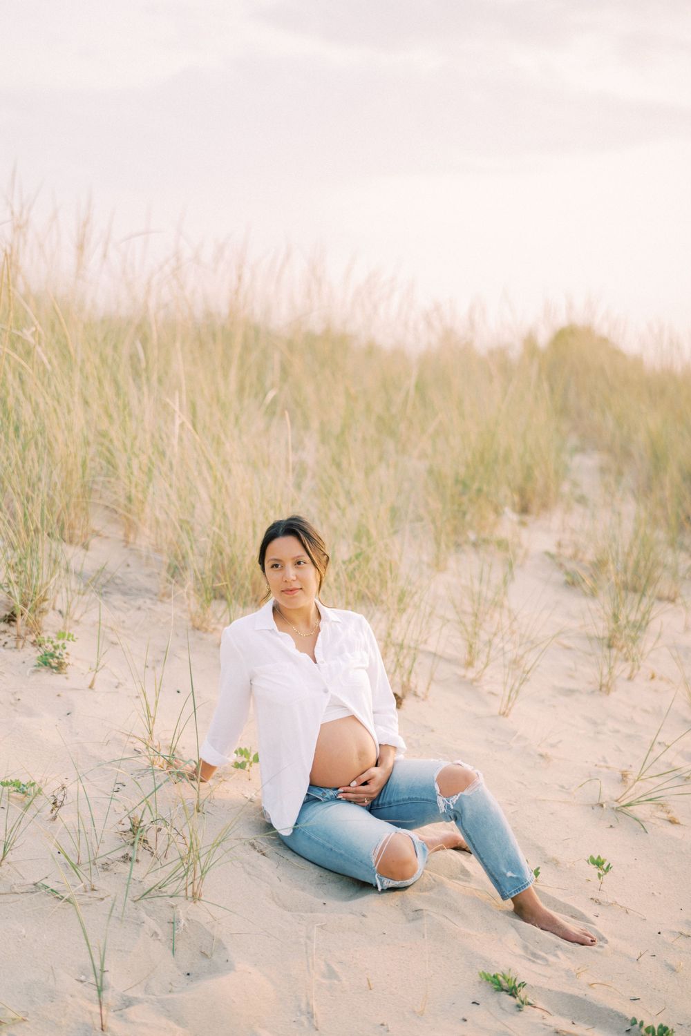 A beachside maternity portrait shows a woman in white and denim relaxing among beach grass.
