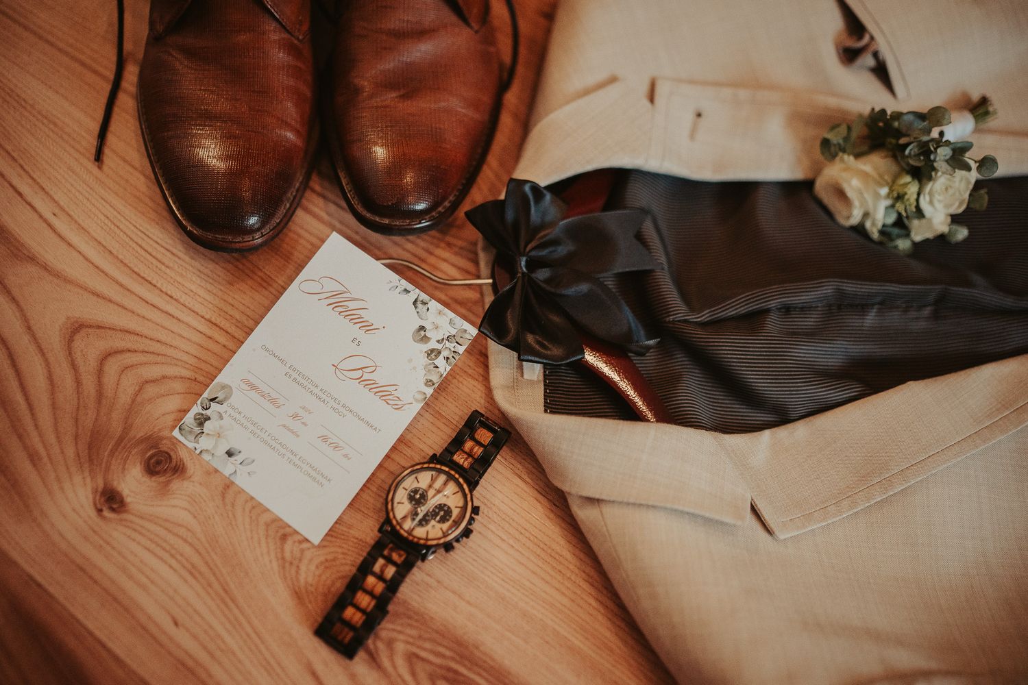 Wedding details including brown leather shoes, watch, boutonniere and invitation card on wooden surface.