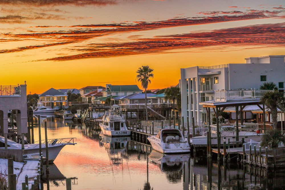 Waterfront marina with boats docked at sunset displaying vibrant orange and pink sky over coastal homes and palm trees.