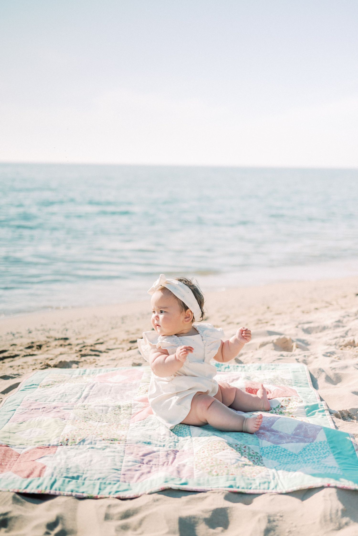 Baby in white dress sits on colorful quilt at beach during sunset.