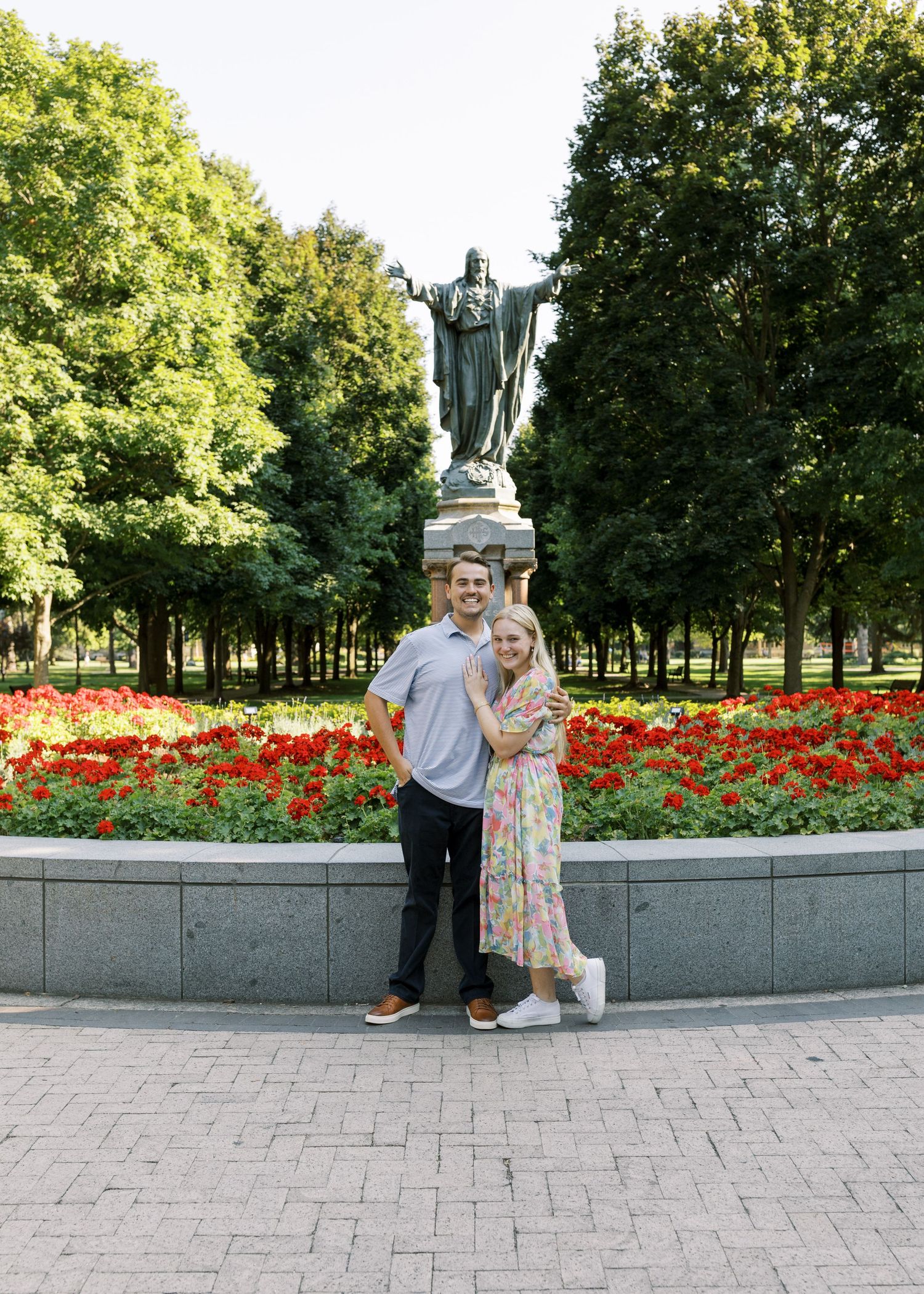 A couple poses in front of a large bronze statue with red flowers blooming in the garden.