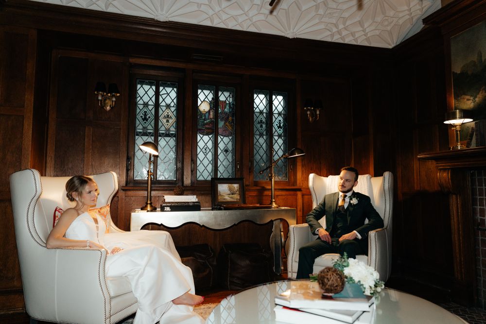 Two people relax together in a luxurious vintage sitting room with wood paneling and stained glass windows.