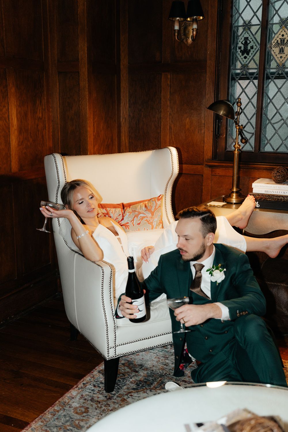 A couple relaxes with drinks in an elegant wood-paneled room with a white armchair and vintage decor.