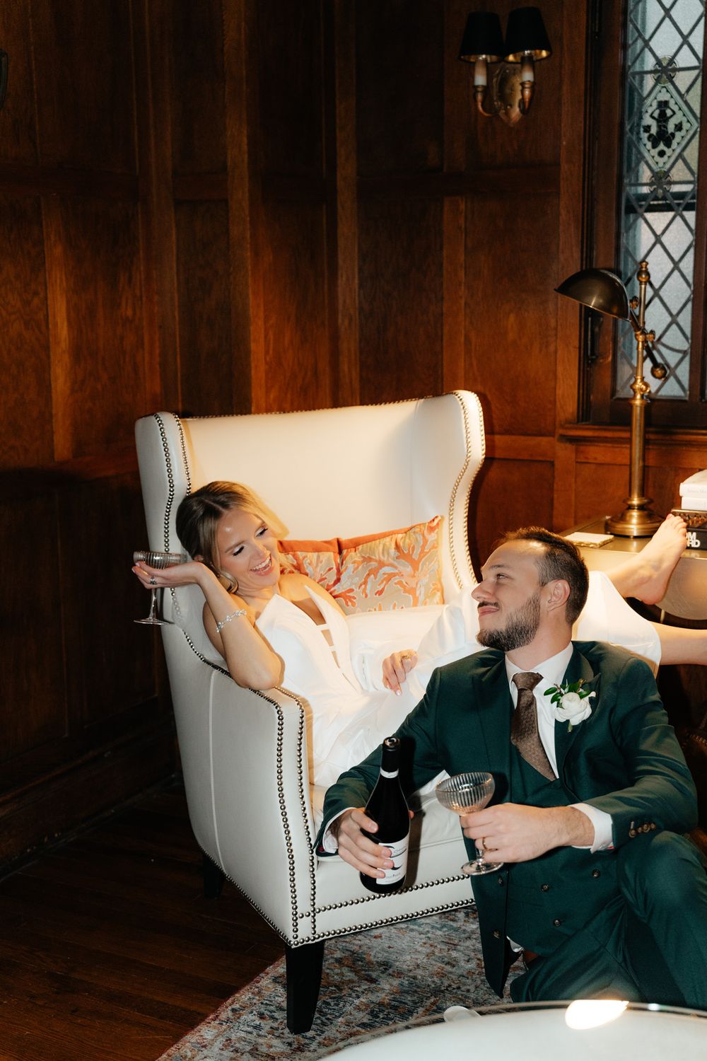 Couple relaxes in a cozy study room with wood paneling as they share drinks in elegant evening attire.