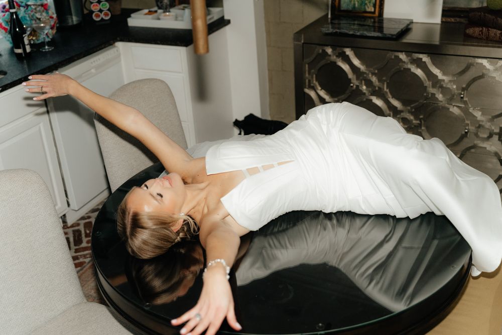 Person in white dress lounges dramatically across a dark circular surface in an interior setting.