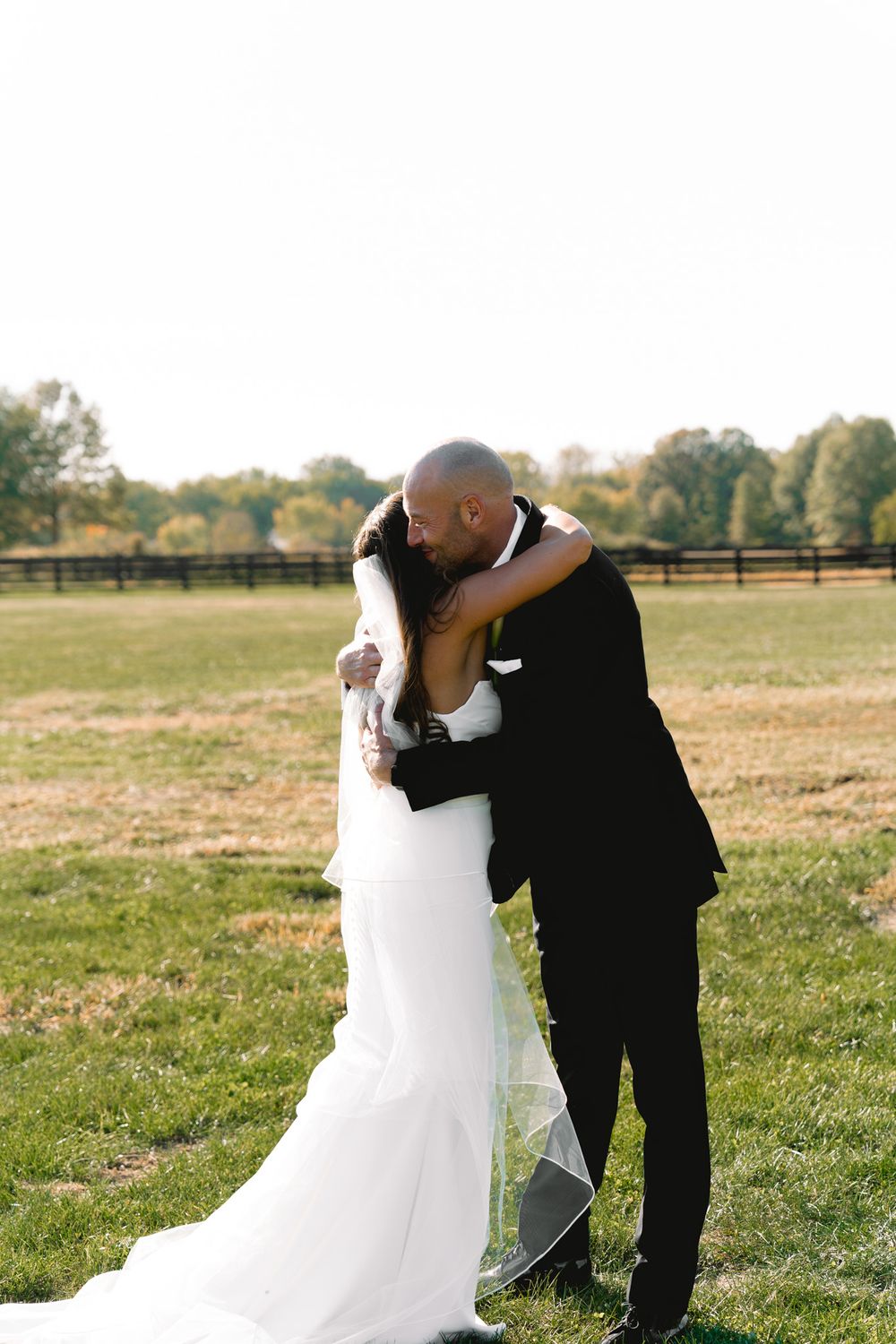 A romantic outdoor wedding moment with a bride in white gown and groom in black suit sharing a tender embrace in a field.