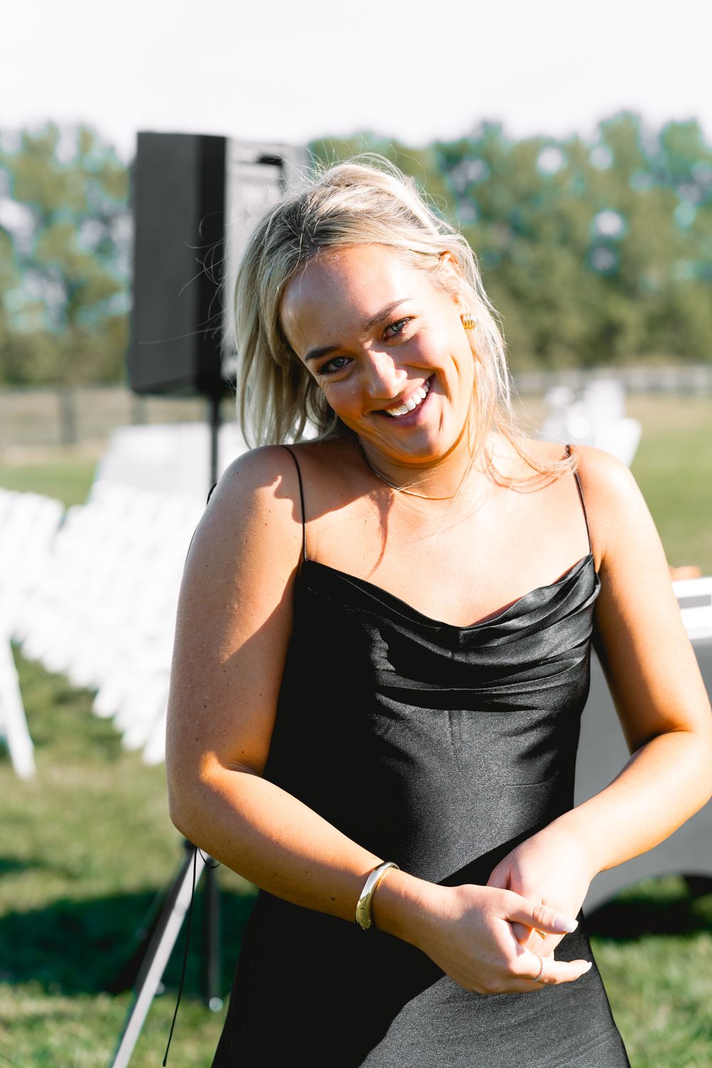 Woman in black dress laughs joyfully outdoors at sunny event with white chairs set up in background.
